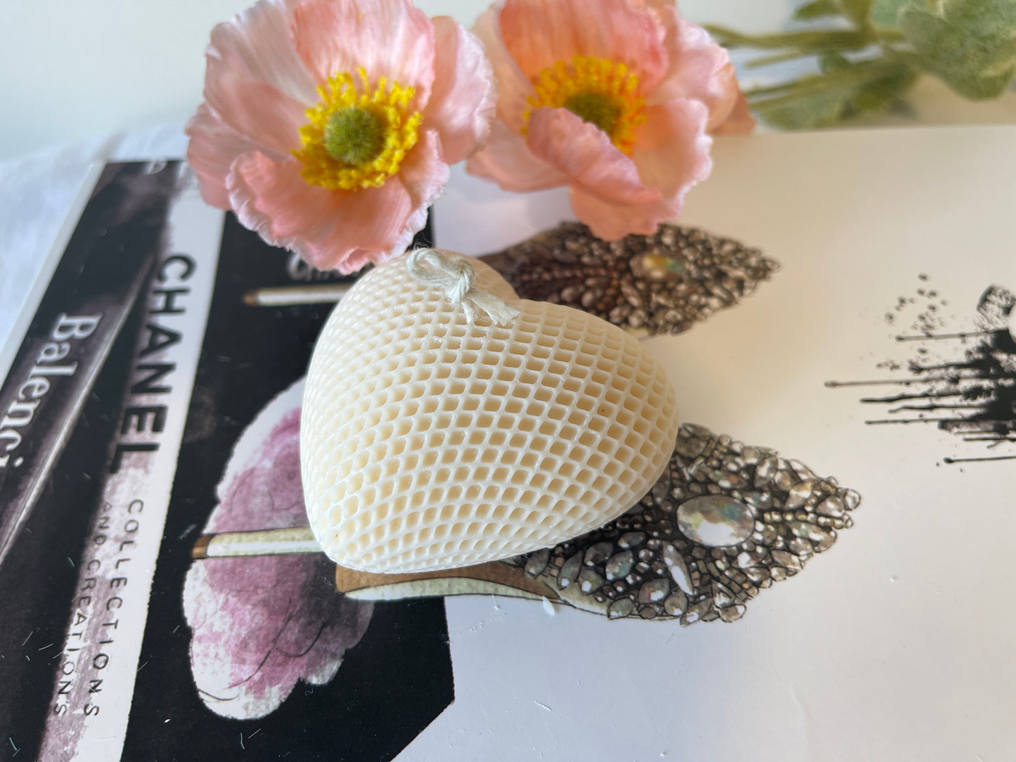 A heart-shaped candle with a textured surface is placed on top of a magazine featuring fashion illustrations, with two soft pink artificial flowers in the background.